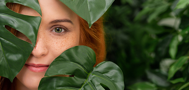 Natural cosmetics and skincare. Young and beautiful redhead woman with freckles in tropical leaves, panorama with free space