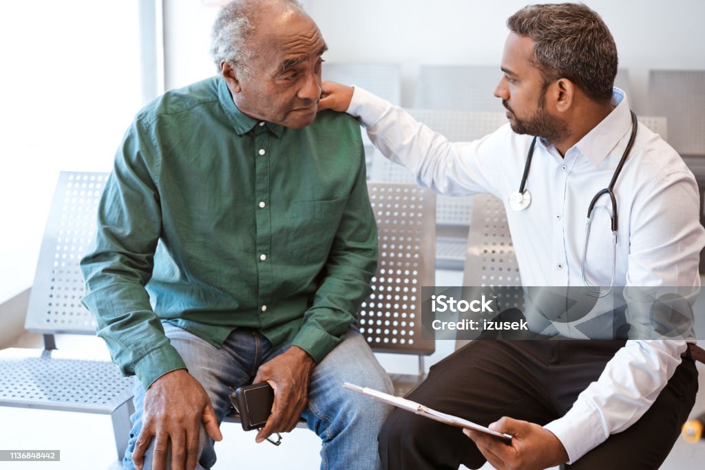 Doctor consoling sad senior male patient Healthcare worker giving bad news with hand on shoulder of male patient. Doctor consoling sad senior man in waiting room. They are sitting at hospital. Doctor Stock Photo