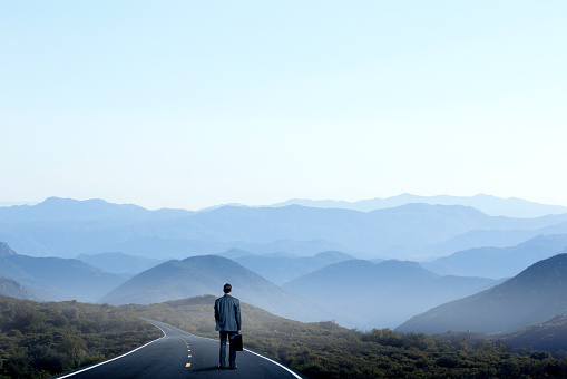 A businessman stands on, and looks down, a long curving rural road that leads toward a series of mountain ridges in the distance.  The scene evokes an emptiness and loneliness that is magnified by the stark and barren nature of the surroundings.