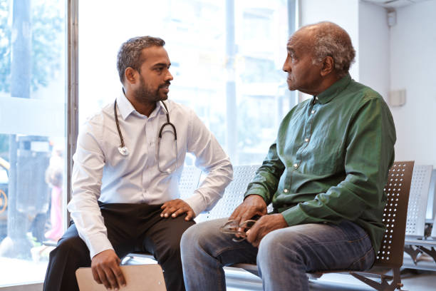 Serious man is staring at doctor in waiting room Doctor talking to senior patient sitting in waiting room. Serious man is staring at healthcare worker at hospital. They are against window. three quarter view stock pictures, royalty-free photos & images