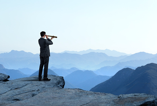 A businessman looks through a spyglass as he stands and looks out towards a series of mountain ridges that recede into the distance