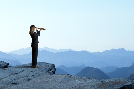 A businesswoman looks through a spyglass as she stands and looks out towards a series of mountain ridges that recede into the distance