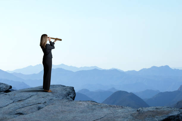 Businesswoman With Spyglass Looking Out Toward Mountain Range A businesswoman looks through a spyglass as she stands and looks out towards a series of mountain ridges that recede into the distance looking through an object stock pictures, royalty-free photos & images