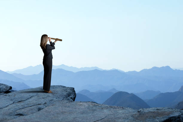 businesswoman with spyglass looking out toward mountain range - olhando através imagens e fotografias de stock