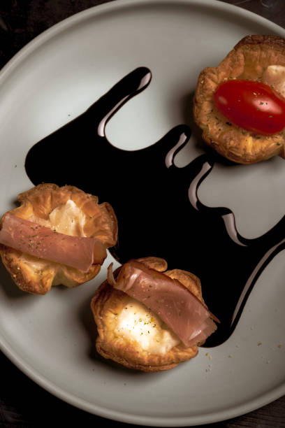 Vertical photo with top view on blue plate with puff pastry cups. Cups are stuffed by melted cheese. Ham and red tomatoes are on baked pastries. Dark sauce is spilled on plate.