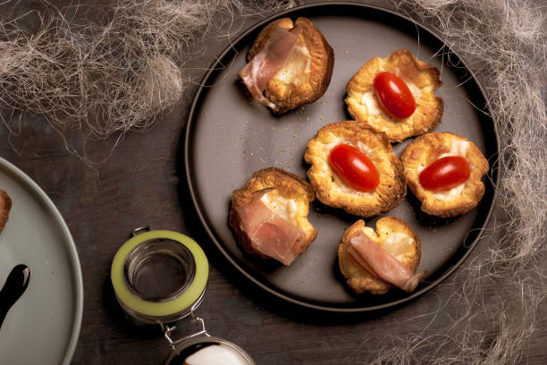 Horizontal photo with top view on black plate with several baked puff pastry cups. Cups are stuffed by cheese. Red tomatoes and dried ham is on pastries. Plate is on dark wooden board.