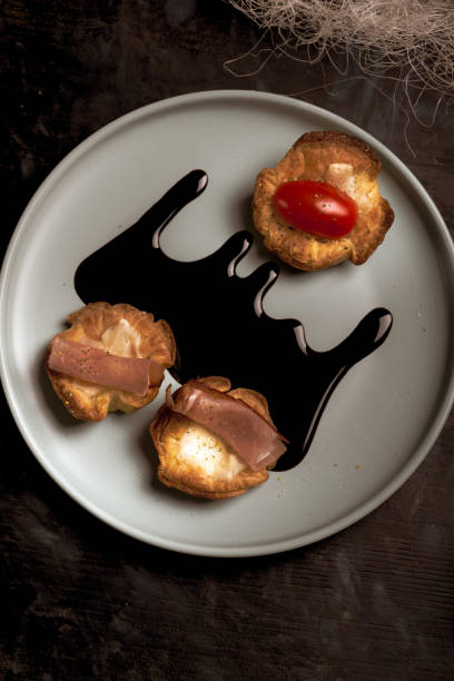 Vertical photo with top view on blue plate with puff pastry cups. Cups are stuffed by melted cheese. Ham and red tomatoes are on baked pastries. Dark sauce is spilled on plate.