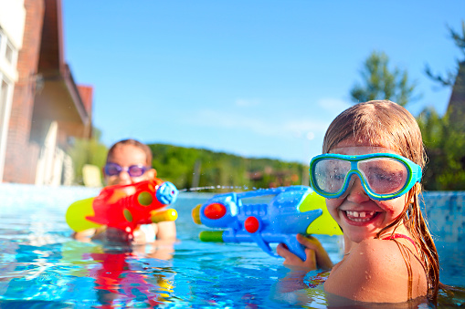 Funny little girls playing with water guns wearing pool masks on summer day in the pool