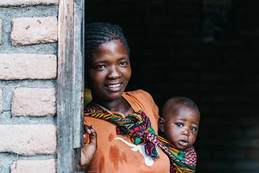 young african mother with baby looking out of the door of typical brick house