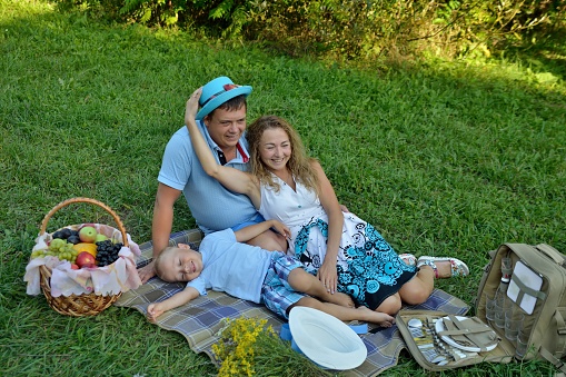 Happy family on a picnic in nature. Mom, dad and son have fun and play lying on the grass in the Park in the summer at sunset near the fruit basket. Portrait. Horizontal orientation of the image.