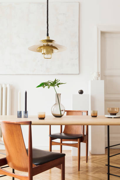 istock Eclectic and elegant dining room interior with design sharing table, chairs, gold pedant lamp, abstract paintings and stylish accessories. Tropical leafs in vase. Eclectic decor. Brown wooden parquet. 1136893811
