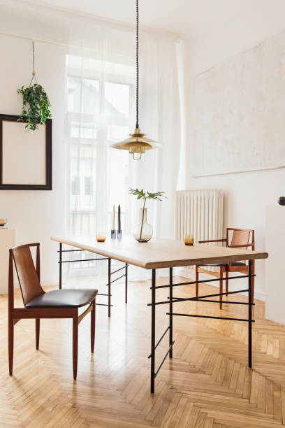 istock Sunny and elegant dining room interior with design sharing table, chairs, gold pedant lamp, abstract paintings and stylish accessories. Tropical leafs in vase. Eclectic decor. Brown wooden parquet. 1136894481