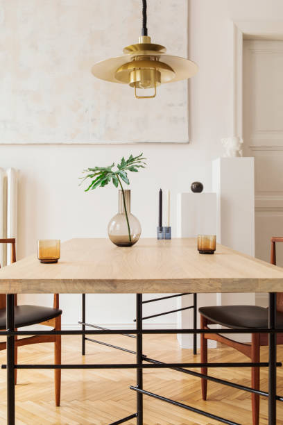 istock Eclectic and elegant dining room interior with design sharing table, chairs, gold pedant lamp, abstract paintings and stylish accessories. Tropical leafs in vase. Eclectic decor. Brown wooden parquet. 1136894563