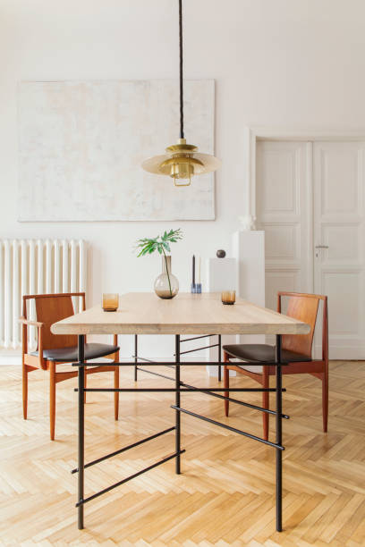 istock Eclectic and elegant dining room interior with design sharing table, chairs, gold pedant lamp, abstract paintings and stylish accessories. Tropical leafs in vase. Eclectic decor. Brown wooden parquet. 1136894588