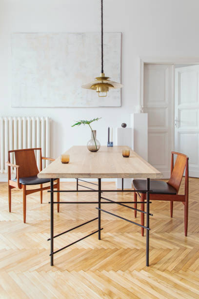 istock Eclectic and elegant dining room interior with design sharing table, chairs, gold pedant lamp, abstract paintings and stylish accessories. Tropical leafs in vase. Eclectic decor. Brown wooden parquet. 1136894595