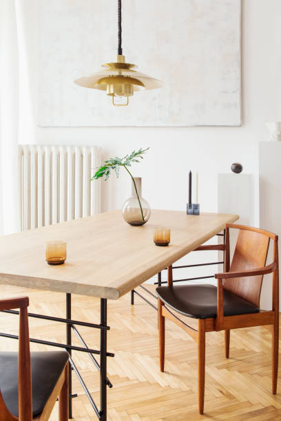 istock Stylish and modern dining room interior with design sharing table, chairs, gold pedant lamp, abstract paintings and elegant accessories. Tropical leafs in vase. Eclectic decor. Brown wooden parquet. 1136894705