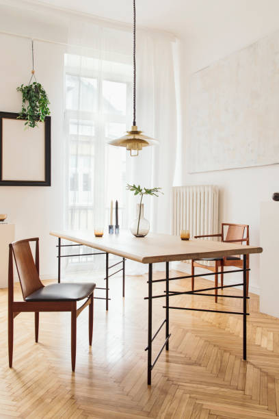 istock Stylish and modern dining room interior with design sharing table, chairs, gold pedant lamp, abstract paintings and elegant accessories. Tropical leafs in vase. Eclectic decor. Brown wooden parquet. 1136894728