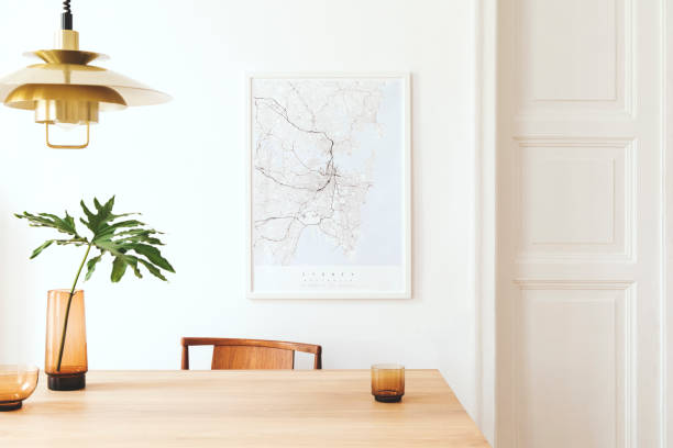 istock Stylish and modern dining room interior with mock up poster map, sharing table design chairs, gold pedant lamp and cups of coffee. White walls, wooden parquet. Tropical leafs in vase. Eclectic decor. 1136895102