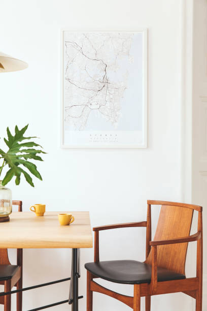 istock Stylish and modern dining room interior with mock up poster map, sharing table design chairs, gold pedant lamp and cups of coffee. White walls, wooden parquet. Tropical leafs in vase. Eclectic decor. 1136895193