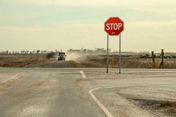 landstraßen-stoppschild mit großem einschussloch steht an kreuzung mit pickup-truck mit anhänger, der in einer staubwolke auf ihn zufährt - oklahoma sign road sign sky stock-fotos und bilder