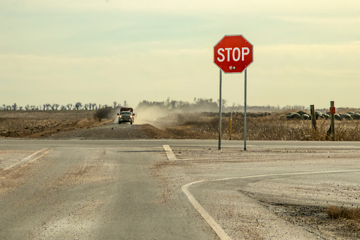 Country Roads - Stop sign with large bullet hole stands by crossroads with pickup truck with trailer driving toward it in a cloud of dust