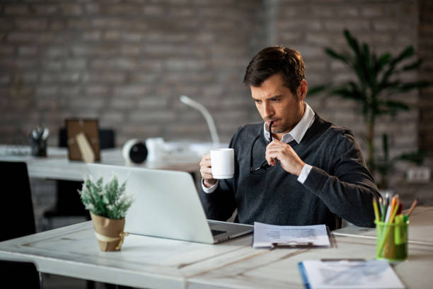 Pensive businessman working on laptop while drinking coffee in the office. Mid adult businessman using computer while working on reports and drinking coffee at work. mid adult men stock pictures, royalty-free photos & images