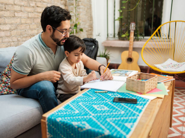 istock Mexican father sitting with boy at home and writing on notebook 1136937773