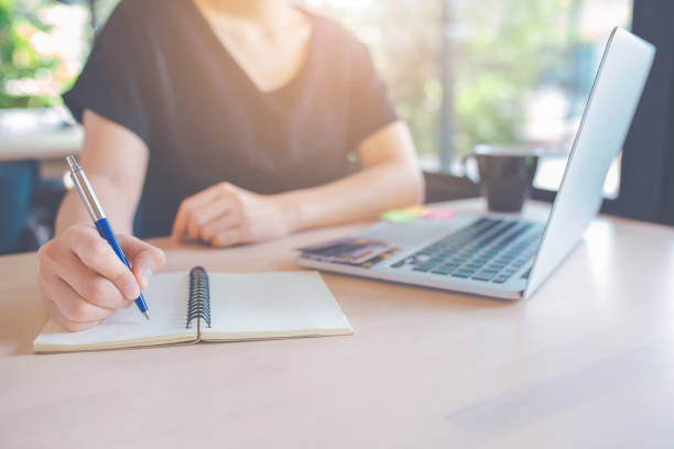 istock Woman hand is writing on a notepad with a pen.On the table, there is a laptop computer and a cup of coffee. 1136945012
