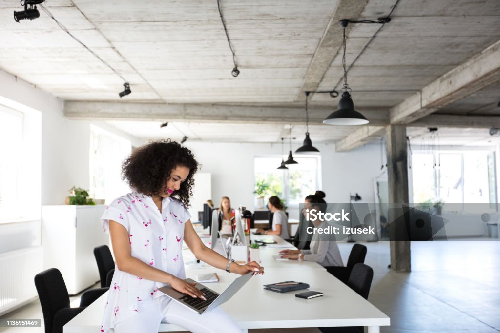 Young woman with laptop at office Young woman with laptop sitting at the table with coworkers working on computers in background. Computer Software Stock Photo