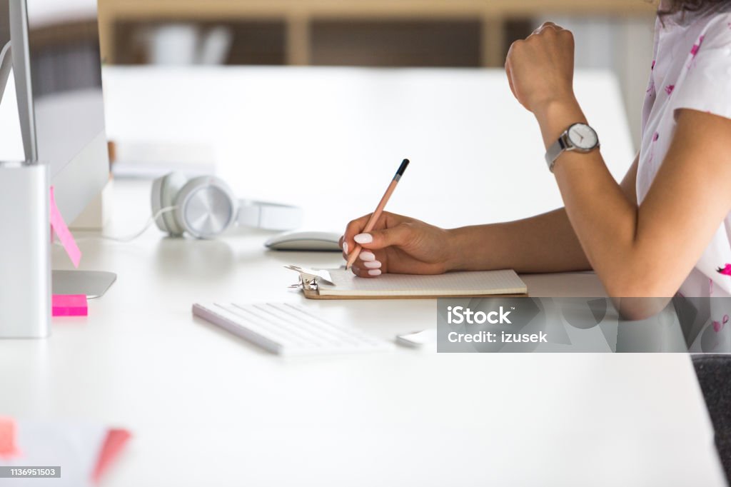 Woman making notes at office desk Close up of female hands writing on notepad while working at her desk. Focus on female hands holding pencil making notes while working in office. Clipboard Stock Photo