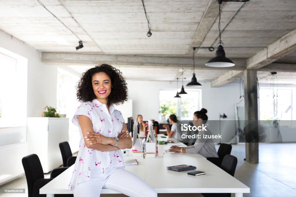 Confident female software engineer in modern office Portrait of young woman sitting at the table with colleagues working on computer in background. Desk Stock Photo