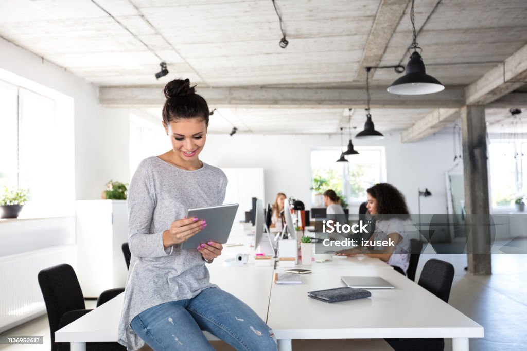 Beautiful young woman with digital tablet at office Beautiful young woman with digital tablet sitting at the table with coworkers working on computers in background. 20-24 Years Stock Photo