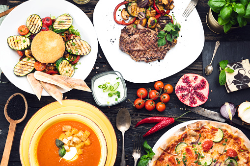 Grilled steak with grilled vegetables, salad, soup and pizza  on a dark wooden table, top view