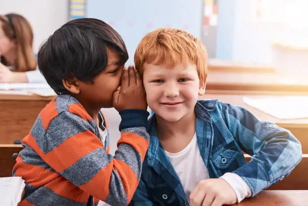Cropped shot of elementary school kid whispering into another student's ear inside of the classroom