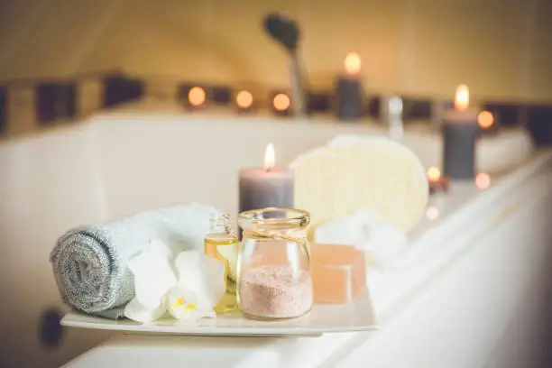Photo of White ceramic tray with home spa supplies in home bathroom for relaxing rituals. Candlelight, salt soap bar, bath salt in jar, massage, bath oil in bottle, blue rolled towel, natural sponge.