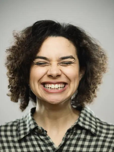 Close-up portrait of young caucasian happy woman with excited expression and eyes closed. Vertical shot of hispanic real people laughing with long curly hair and green eyes. Photography from a DSLR camera. Sharp focus on eyes.
