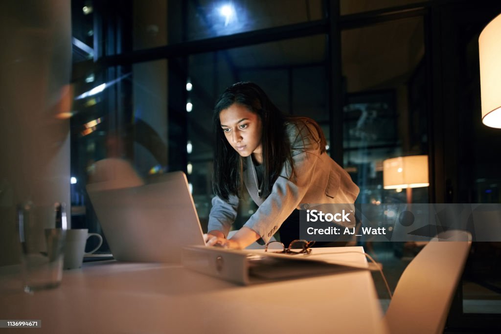 Downloading the last file and then I'm done! Shot of a young businesswoman using her laptop while working late at the office Persistence Stock Photo