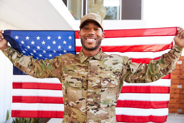 Millennial black soldier standing outside modern building holding US flag, smiling to camera, close up Millennial black soldier standing outside modern building holding US flag, smiling to camera, close up black military man stock pictures, royalty-free photos & images