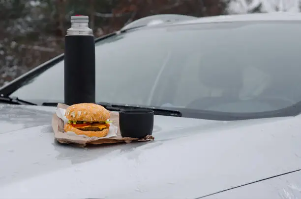 Photo of Winter picnic with tea and burger on the hood of a silver car against the background of the forest