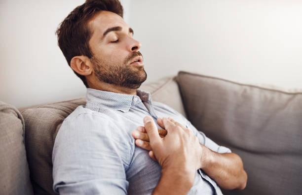 Let's just hope this isn't serious Shot of a handsome young businessman holding his chest in pain while relaxing on a sofa at home male chest pain stock pictures, royalty-free photos & images