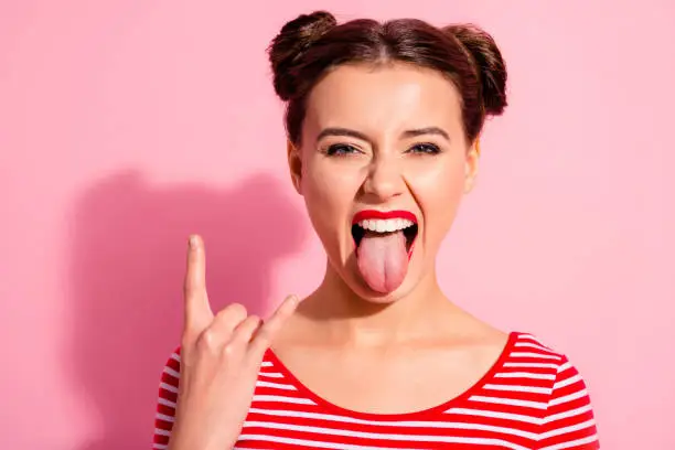 Close up photo beautiful carefree she her lady pretty hairdo two buns bright red pomade big lips tongue out mouth rocker gesture wear casual striped red white t-shirt isolated pink background.