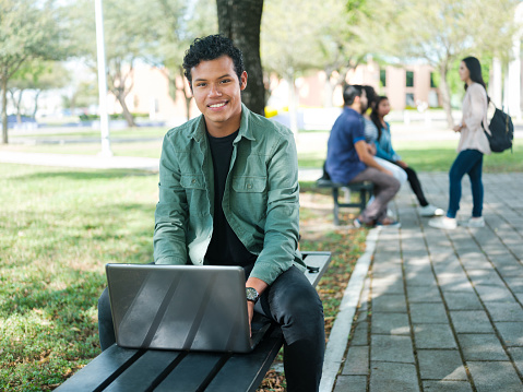 A young confident male university student sitting on a bench with a laptop looking at the camera outside