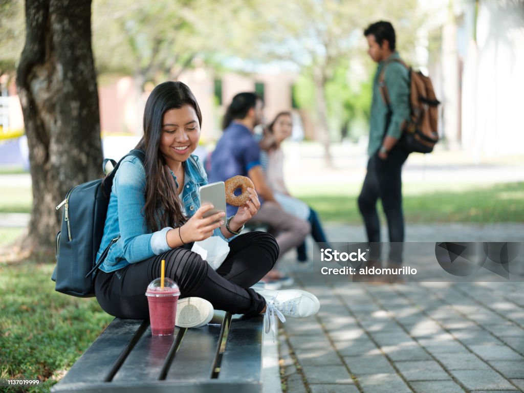 College student snacking and texting A latin college girl snacking and texting while sitting on a bench. Student Stock Photo