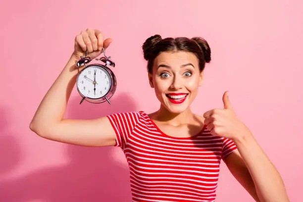 Close-up portrait of her she nice cute charming winsome attractive glamorous cheerful girl wearing striped t-shirt holding in hand showing clock thumbup hour isolated over pink pastel background.