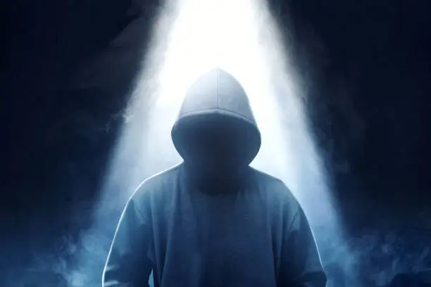 Hacker in black hoodie standing with smoke and light from the top over dark background