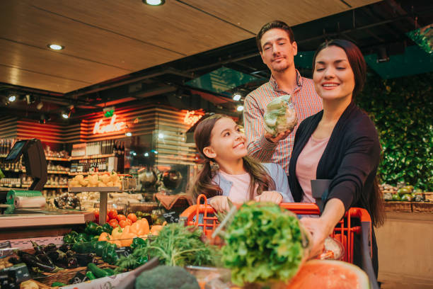 young parents and daughter in grocery store. father hold cauliflower in hands. mother has lettuce and look at it. daughter smile. she looks at mother. - leaf vegetable freshness vegetable market imagens e fotografias de stock