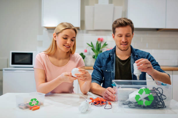 リサイクル、再利用、エネルギー。若い家族は台所で座っている間リサイクルの記号が付いている容器に電球、電池、他の電子廃棄物を分類します - recycled bulb ストックフォトと画像