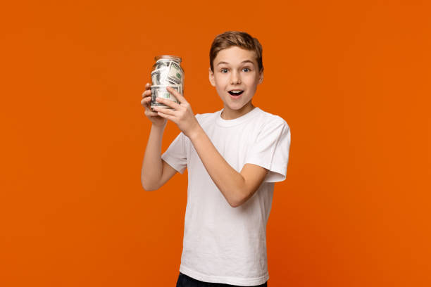 emocjonalny chłopiec trzymający szklaną skrzynkę z pieniędzmi na pomarańczowym tle - economize zdjęcia i obrazy z banku zdjęć