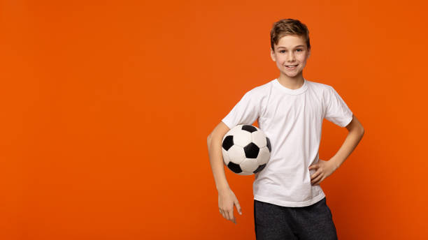 garçon posant avec le ballon de football sur le fond de studio orange - anticipation smiling touching image technique photos et images de collection