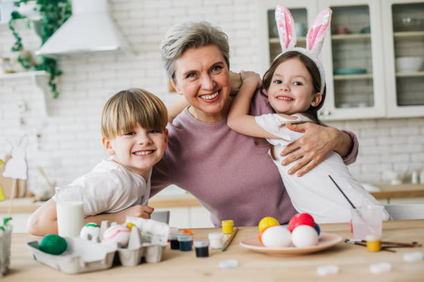 mezza lunghezza di donna dai capelli grigi che abbraccia i suoi nipoti in cucina - easter easter egg paintbrush holding foto e immagini stock